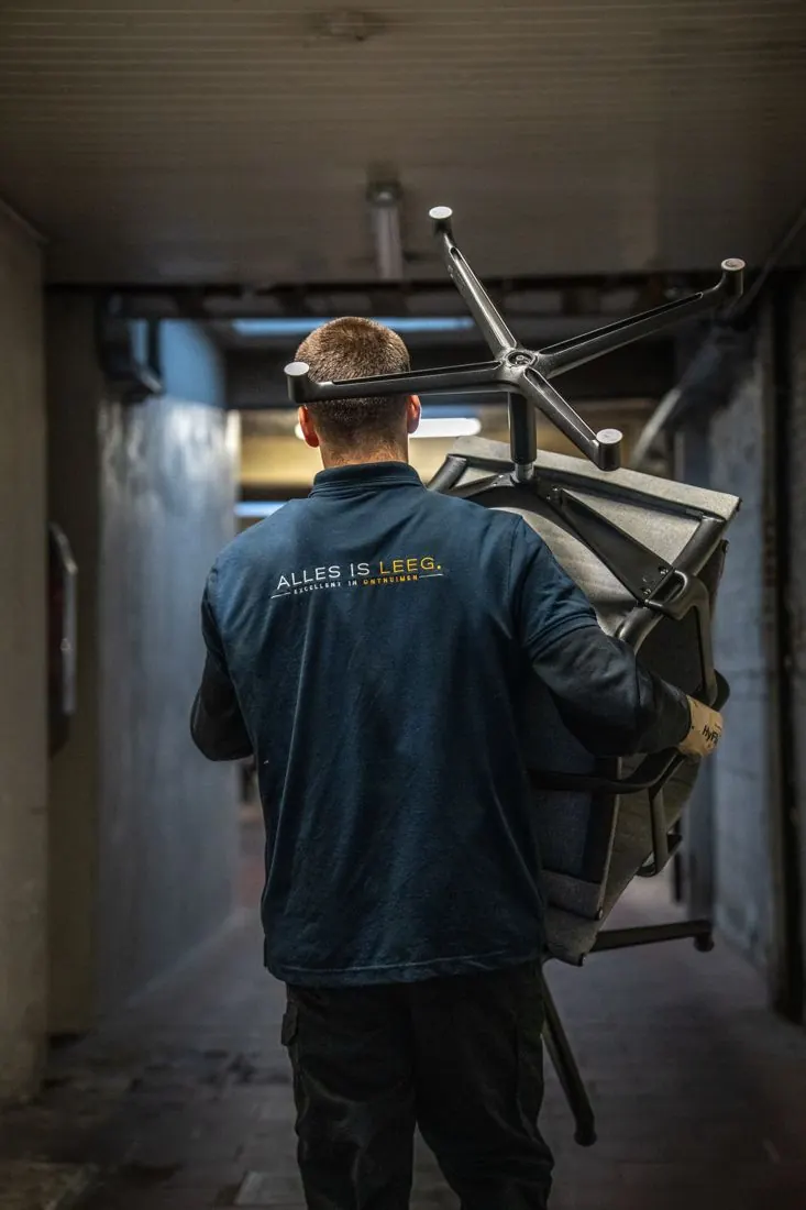 Medewerker van Alles Is Leeg draagt een bureaustoel door een gang