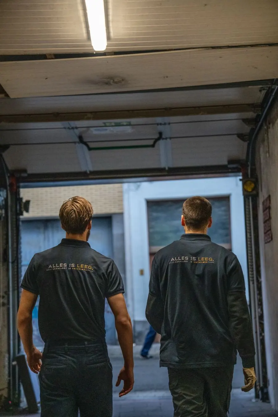 Twee medewerkers van Alles Is Leeg in uniform stappen een garage binnen
